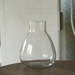 Könnte beinhalten: Eine Vase aus klarem Glas mit einem breiten, abgerundeten Boden, der sich nach oben hin zu einem schmaleren, zylindrischen Hals verjüngt. Das Gefäß besitzt eine glatte, transparente Oberfläche und einen einfachen, offenen oberen Rand.