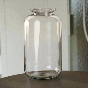Könnte beinhalten: Eine hohe, zylindrische Glasvase mit glatter, transparenter Oberfläche und einem leicht ausgestellten, abgerundeten Rand. Das Gefäß hat einen breiten, abgerundeten Körper, der sich nach oben hin sanft verjüngt und eine klare, schlichte Form bildet.