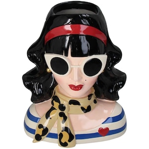 Puede incluir: Un jarrón de cerámica con forma de busto de mujer. La mujer tiene el pelo negro, labios rojos y lleva gafas de sol blancas, una bufanda de estampado de leopardo y una camisa a rayas azules y blancas.