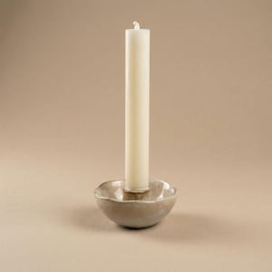 Könnte beinhalten: Eine cremefarbene Säulenkerze steht in einem kleinen, runden Kerzenhalter aus Keramik. Der Kerzenhalter hat eine strukturierte, organische Form mit hellbrauner Glasur. Der Hintergrund ist einfarbig, neutral beige.