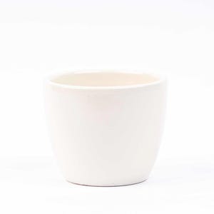 Op de afbeelding: Een witte keramische pot met een afgeronde vorm. De pot is leeg en klaar voor een plant.