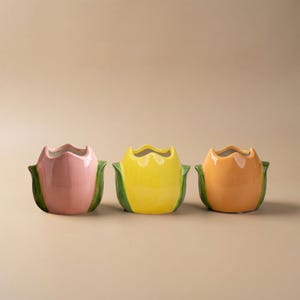 Könnte beinhalten: Drei tulpenförmige Keramikvasen. Die Vasen sind in Rosa, Gelb und Orange gehalten, mit grünen Basen. Die Vasen haben eine glänzende Oberfläche. Jede Vase ist etwa 10 cm hoch und leer.