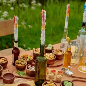 Peut inclure: Une table dressée avec diverses collations et boissons, dont une bouteille de vin dans laquelle est insérée une bougie colorée. La table est décorée de bols et d'assiettes en bois, et il y a plusieurs autres bougies sur la table.