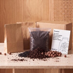 Op de afbeelding: Een terrariumset met twee bruine papieren zakken, een doorzichtige plastic zak gevuld met donkere potgrond, een kartonnen doos en een witte handleiding met de tekst Terrarium, Gebruikershandleiding en een lijntekening van een plant.