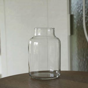 May include: A clear glass vase with a cylindrical body and a slightly tapered neck. The vase features a smooth, transparent surface and a wide, circular opening at the top, showcasing a simple, functional design for holding various floral arrangements.