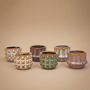 Può includere: Una collezione di sei piccoli vasi in ceramica di vari colori. I vasi hanno motivi geometrici strutturati e una finitura lucida. I colori includono azzurro, marrone, beige, verde, grigio e viola. I vasi sono disposti su uno sfondo neutro.