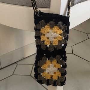 Puede incluir: Una bolsa de crochet negra con un patrón de granny square amarillo y blanco. La bolsa tiene una correa larga hecha de hilo negro y amarillo.