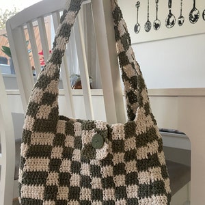 Könnte beinhalten: Eine grün-weiß karierte gehäkelte Tasche mit einem einzigen Knopfverschluss. Die Tasche hängt an einem weißen Stuhl.