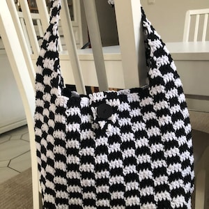 Könnte beinhalten: Handgefertigte Häkeltasche mit schwarz-weißem Schachbrettmuster. Die Tasche hat einen großen schwarzen Knopf und einen langen Schulterriemen. Die Tasche liegt auf einem weißen Stuhl. Ungefähre Abmessungen: 30 cm breit und 30 cm hoch.