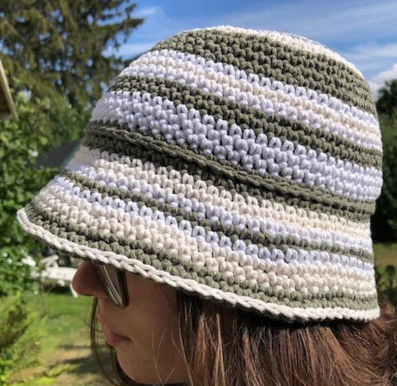 Puede incluir: Un sombrero de cubo de crochet a rayas verdes y blancas. El sombrero tiene un ala ancha y est&aacute; hecho con un patr&oacute;n de punto simple.