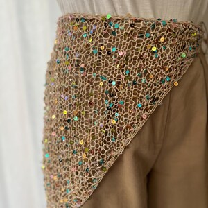 May include: A beige crocheted belt adorned with colorful sequins. The belt is triangular in shape and appears to be worn around the waist. The sequins are in various colors, including blue, green, pink, and gold.