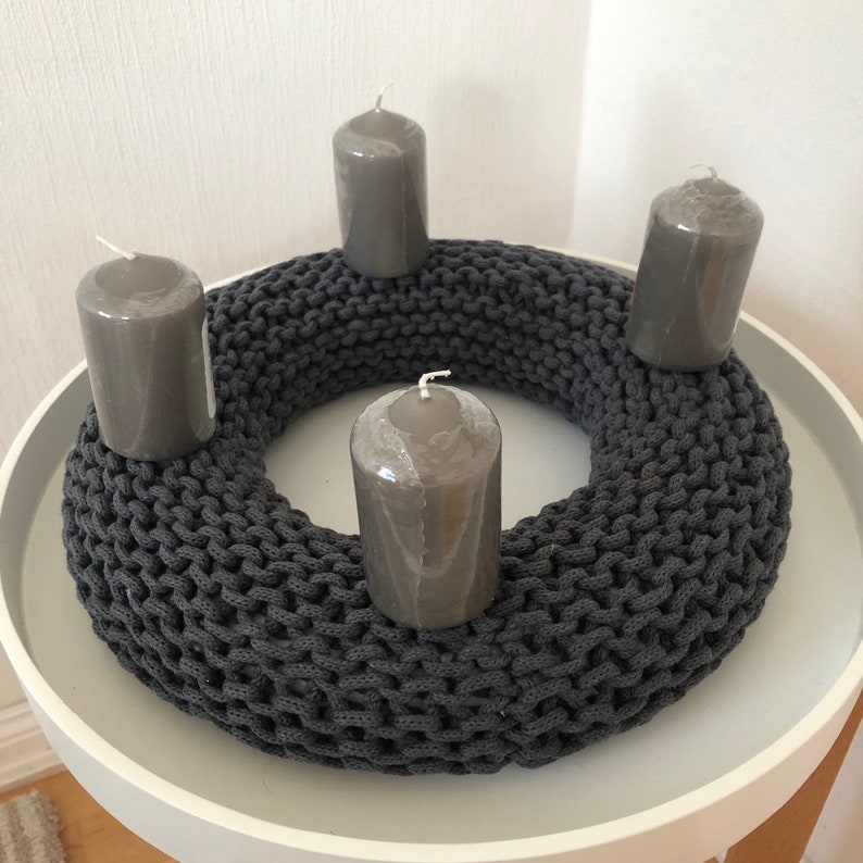May include: A gray knitted Advent wreath with four gray candles. The wreath is made of thick, chunky yarn and has a simple, modern design.