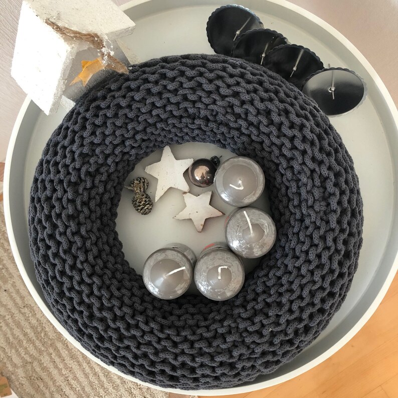 May include: A gray knitted wreath with four white candles and a silver ornament. The wreath is on a white plate with a white background.