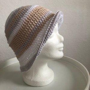 Könnte beinhalten: Eine gehäkelte Mütze mit weißen, beigen und grauen Streifen. Die Mütze hat einen breiten Rand und wird auf einem weißen Mannequin-Kopf präsentiert.
