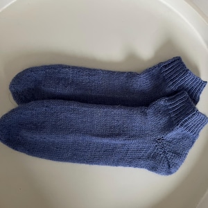 Op de afbeelding: Een paar blauwe gebreide sokken. De sokken zijn effen van kleur en lijken gemaakt van een zacht, getextureerd materiaal. De sokken liggen op een wit oppervlak.