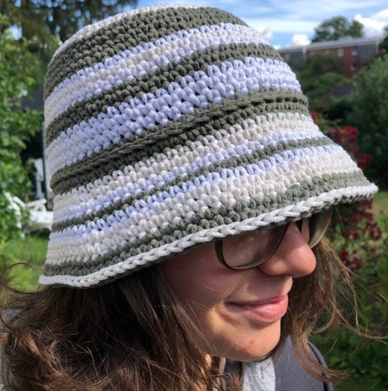 Puede incluir: Un sombrero de cubo de crochet a rayas verdes y blancas. El sombrero tiene un ala ancha y est&aacute; hecho con un hilo suave y texturizado.