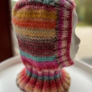 Könnte beinhalten: Handgestrickte Sturmhaube in leuchtenden Farben wie Pink, Orange, Gelb und Türkis. Die Sturmhaube hat einen gerippten Kragen und ein gestreiftes Muster. Sie bedeckt Kopf und Hals.