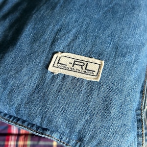 Könnte beinhalten: Nahaufnahme eines blauen Denim-Stoffs mit einem genähten Saum. Ein rechteckiges Etikett mit dem Text "L-RL LAUREN RALPH LAUREN" ist auf den Stoff genäht. Der Denim hat ein strukturiertes Aussehen.