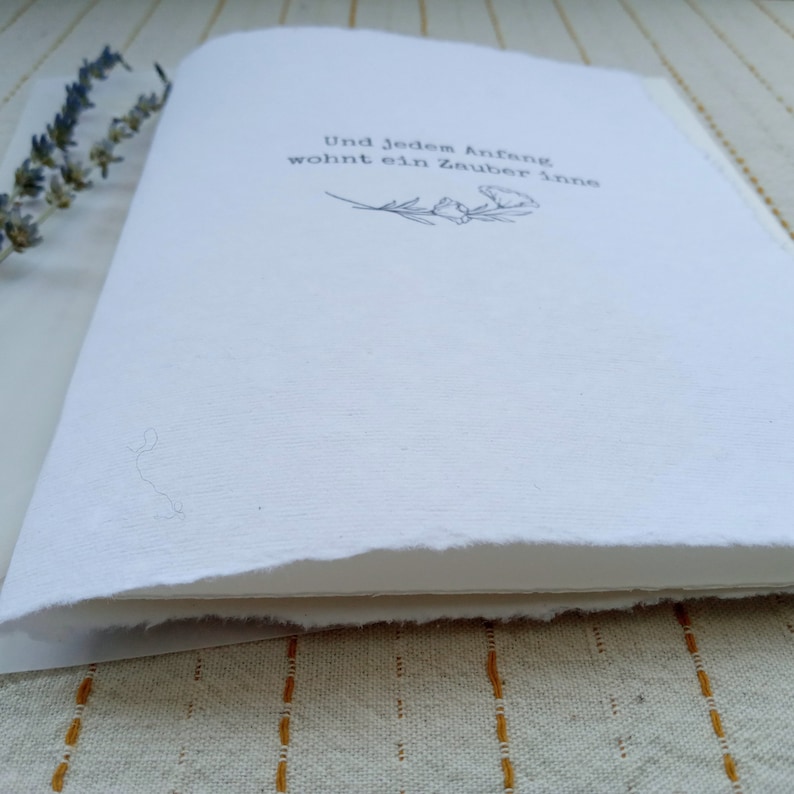 Może przedstawiać: Biała książka z prostym wzorem kwiatowym i tekstem "Und jedem Anfang wohnt ein Zauber inne" nadrukowanym na okładce.