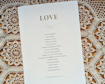 May include: A rectangular white paper print featuring the word LOVE in large capital letters at the top, followed by the text of 1 Corinthians 13:4-7 in a dark grey serif font, centered with a small decorative sprig graphic below the title.