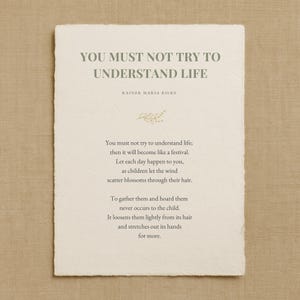 May include: An off-white rectangular card with a deckled edge displays the quote "YOU MUST NOT TRY TO UNDERSTAND LIFE" by Rainer Maria Rilke. A poem about life is printed below the quote, with a small floral illustration.