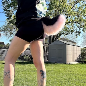 以下が含まれることがあります： 黒とピンクのフェイクファーの尻尾が黒いショートパンツに付いています。 尻尾はふわふわで、黒いベースとピンクの先端が付いています。 人は、小屋と木々の前の緑の草の上に立っています。