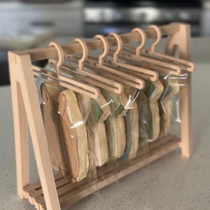 Può includere: Un espositore in legno con grucce in miniatura che contengono biscotti confezionati a forma di camicia. I biscotti sono di vari colori, tra cui beige, verde e blu. L'espositore è in legno chiaro e poggia su un piano di lavoro.