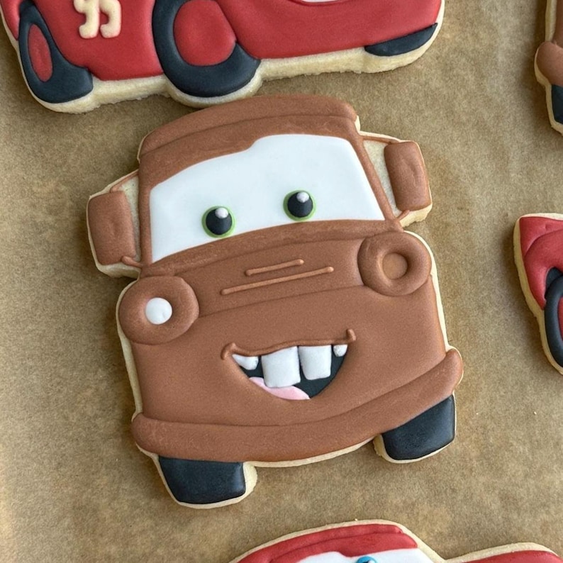 May include: Brown cookie shaped like the character Mater from the movie Cars, with white, black, and green details. The cookie has a smiling face and is surrounded by other car-shaped cookies.