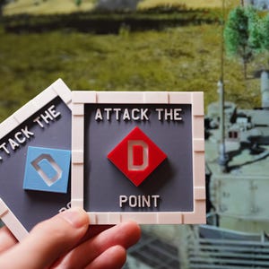 Könnte beinhalten: Zwei quadratische Spielsteine mit dem Text "ATTACK THE POINT". Ein Stein hat eine rote Raute mit einem weißen "D", der andere hat ein blaues Quadrat mit einem weißen "D". Der Hintergrund zeigt eine militärische Szene.