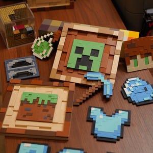May include: A collection of pixelated Minecraft-themed items, including framed images of a Creeper face and a dirt block, a pickaxe, armor, and other game elements. The items are arranged on a wooden surface.