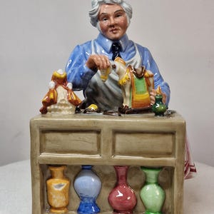 Può includere: Una statuetta in ceramica di un artigiano con camicia blu e grembiule bianco, al lavoro su un banco da lavoro color marrone chiaro. Il banco presenta uno scaffale con quattro piccoli vasi gialli, blu, rossi e verdi. Sul piano ci sono miniature.