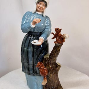 Può includere: Una statuina in porcellana di una persona anziana che indossa una cuffia bianca, una maglia azzurra a maniche lunghe e un grembiule blu scuro. La figura tiene in mano un piattino bianco con delle noci, mentre due scoiattoli marroni sono appollaiati su un tronco d'albero.