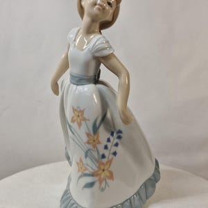 Può includere: Statuina in porcellana di una giovane donna in un abito bianco con accenti floreali gialli e blu. L'abito ha una fascia blu e un orlo a balze. La donna ha i capelli castani e guarda in alto.