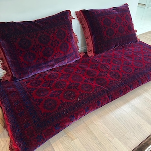 May include: Two red velvet floor cushions with a dark blue and red patterned design and fringe trim. The cushions are arranged side by side on a light wood floor.