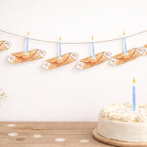 May include: A festive birthday scene featuring a white cake with a single lit blue candle and a garland of watercolor cannoli with blue candles. The garland is strung across a white wall, creating a celebratory atmosphere.
