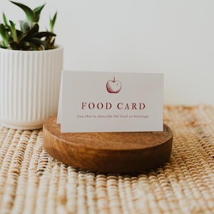 May include: A white food card with a red apple illustration and the words "FOOD CARD". The card is displayed in a round, wooden holder. The text below the words reads "Use this to describe the food or beverage". A white potted plant is in the background.
