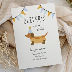 May include: A white birthday invitation card featuring a brown dachshund wearing a blue party hat, decorated with yellow and blue bunting flags. The text reads: OLIVER'S a weenie bit older, bring your buns over, Saturday, October 11th at 2:00, 12 Main Street, City, RSVP to Kate 000.000.0000.