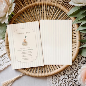 May include: Two cream-coloured invitations with vertical stripes. One features the text "Chapter Two" and "Oliver's Second Birthday" with a teddy bear illustration. The other is blank. Both are on a woven tray.