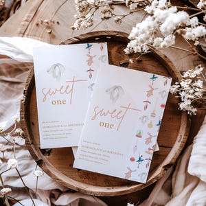 May include: Two white invitation cards with pink and gold lettering that reads "Sweet One" and a watercolor illustration of a snowdrop flower. The cards are on a wooden surface with white flowers.
