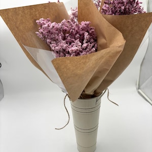 May include: A bouquet of dried pink flowers wrapped in brown paper, presented in a cream-colored metal bucket. The flowers are the focal point, with their delicate texture and vibrant color. The arrangement is tied with twine.