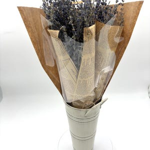 May include: A bouquet of dried lavender flowers wrapped in brown paper and newspaper, displayed in a white metal vase.