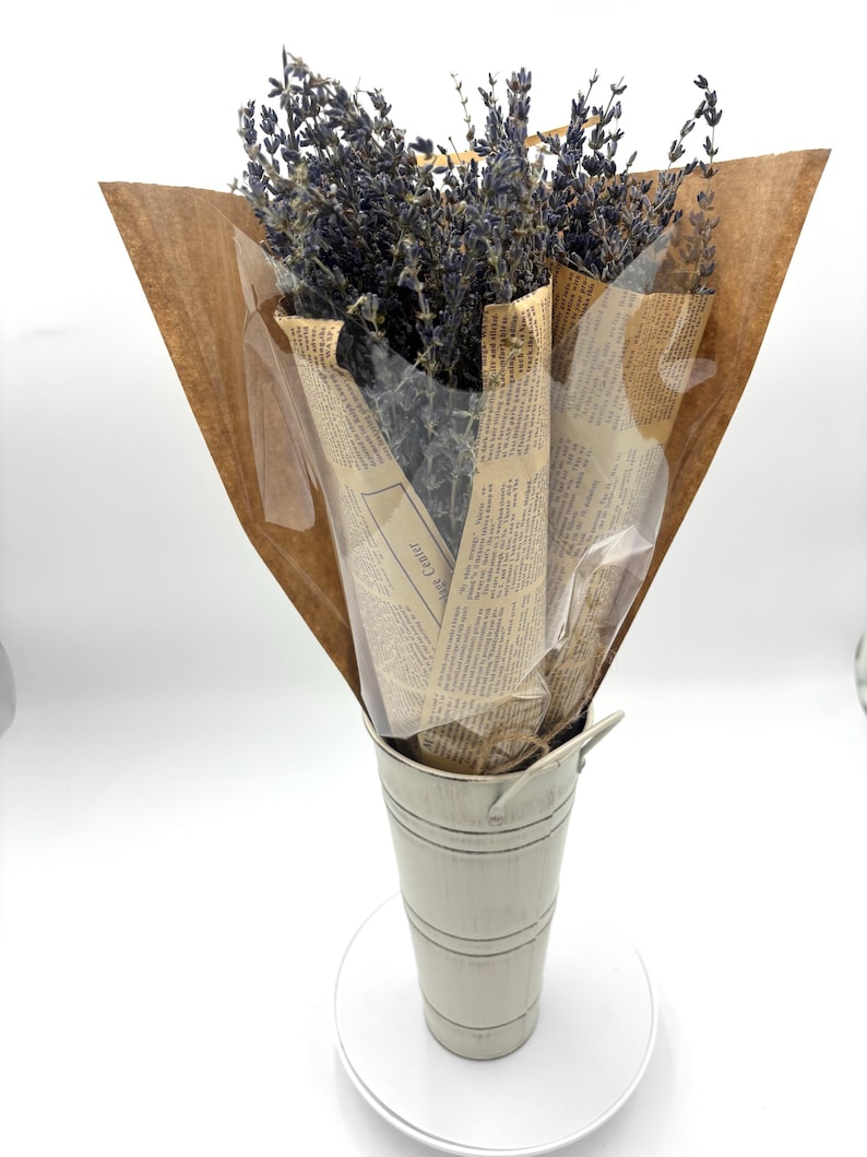 May include: A bouquet of dried lavender flowers wrapped in brown paper and newspaper, displayed in a white metal vase.