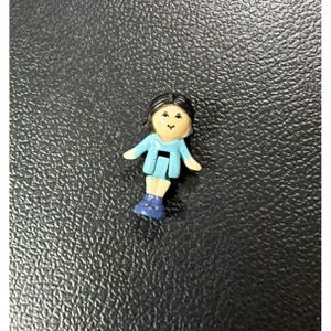 May include: A small plastic doll with black hair, a light skin tone, and a blue outfit. The doll has outstretched arms and blue boots. The doll is a miniature toy figure, likely from a playset or collection.