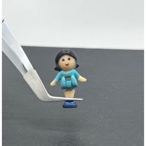May include: A miniature doll figurine with black hair, a light blue dress, and tan arms and legs. The doll is standing on a blue base. The figurine is being held by a pair of silver tweezers.