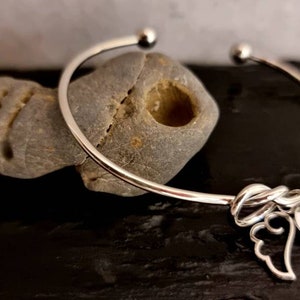 Può includere: Bracciale a cerchio argentato con un design a nodo e due ciondoli: un'ala d'angelo e un piccolo tag rotondo con una lettera. Il bracciale è esposto su una pietra grigia.