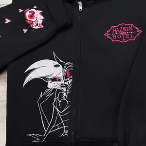 K&ouml;nnte beinhalten: Schwarzer Kapuzenpullover mit Rei&szlig;verschluss, Kapuze und langen &Auml;rmeln. Der linke &Auml;rmel zeigt eine rosa-wei&szlig;e Cartoonfigur und rosa Herzen. Auf der Vorderseite ist eine schwarz-wei&szlig;e Illustration und der Text "HAZBIN HOTEL" in Rosa zu sehen.
