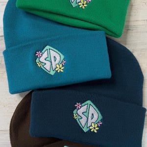 May include: A stack of beanies in various colors: green, teal, navy, and brown. Each beanie features an embroidered patch with the letters "SP" in a diamond shape, surrounded by flowers. The beanies are made of a knitted material.