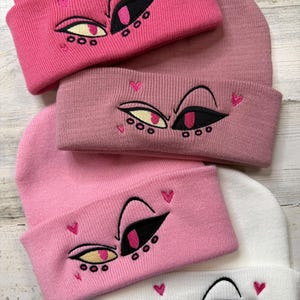 Op de afbeelding: Roze, lichtroze en witte mutsen met geborduurde oogontwerpen. De ogen hebben zwarte, roze en gele details, met kleine roze hartjes. De mutsen zijn in een gebreide stijl, perfect voor koud weer.