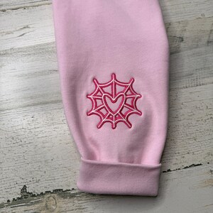 May include: A light pink long-sleeve garment featuring an embroidered design on the sleeve. The embroidery consists of a dark pink spider web pattern surrounding a central dark pink heart outline, finished with a ribbed cuff at the wrist.