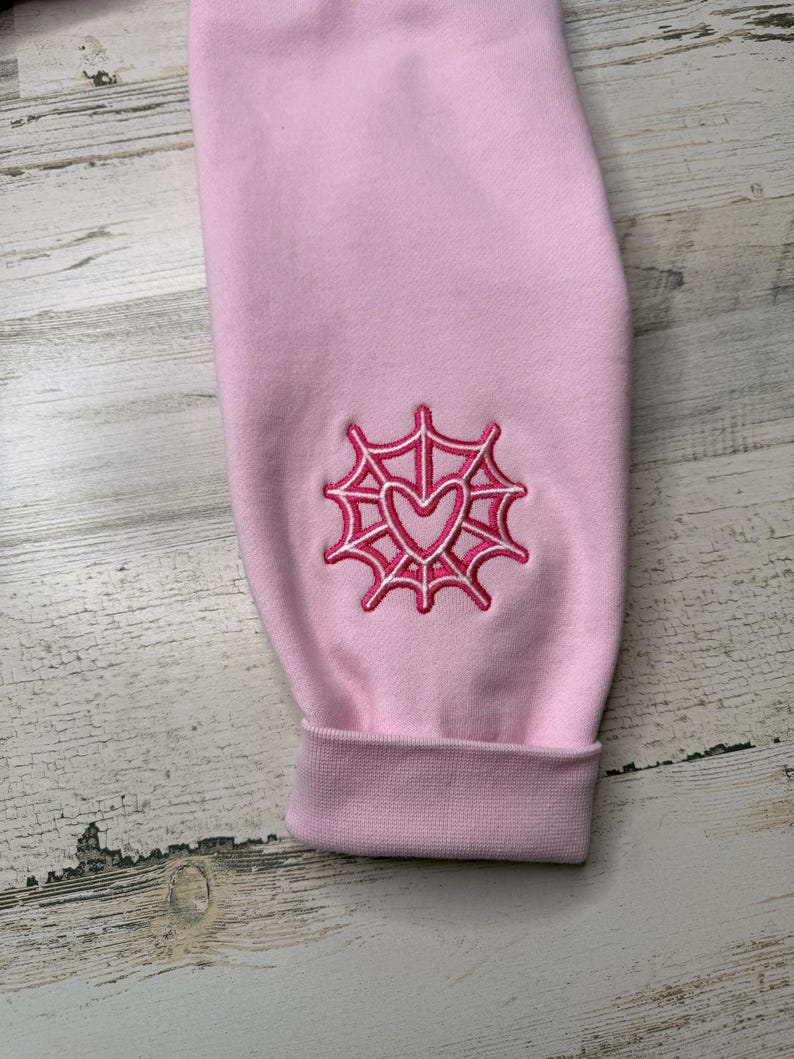 May include: A light pink long-sleeve garment featuring an embroidered design on the sleeve. The embroidery consists of a dark pink spider web pattern surrounding a central dark pink heart outline, finished with a ribbed cuff at the wrist.